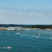 The estuary of the River Hamble leading off the Solent in Hampshire, Southern England, with numerous small boats and yachts manned by unrecognisable people. A marina hosts a forest of yacht masts and the South Downs are in the background..